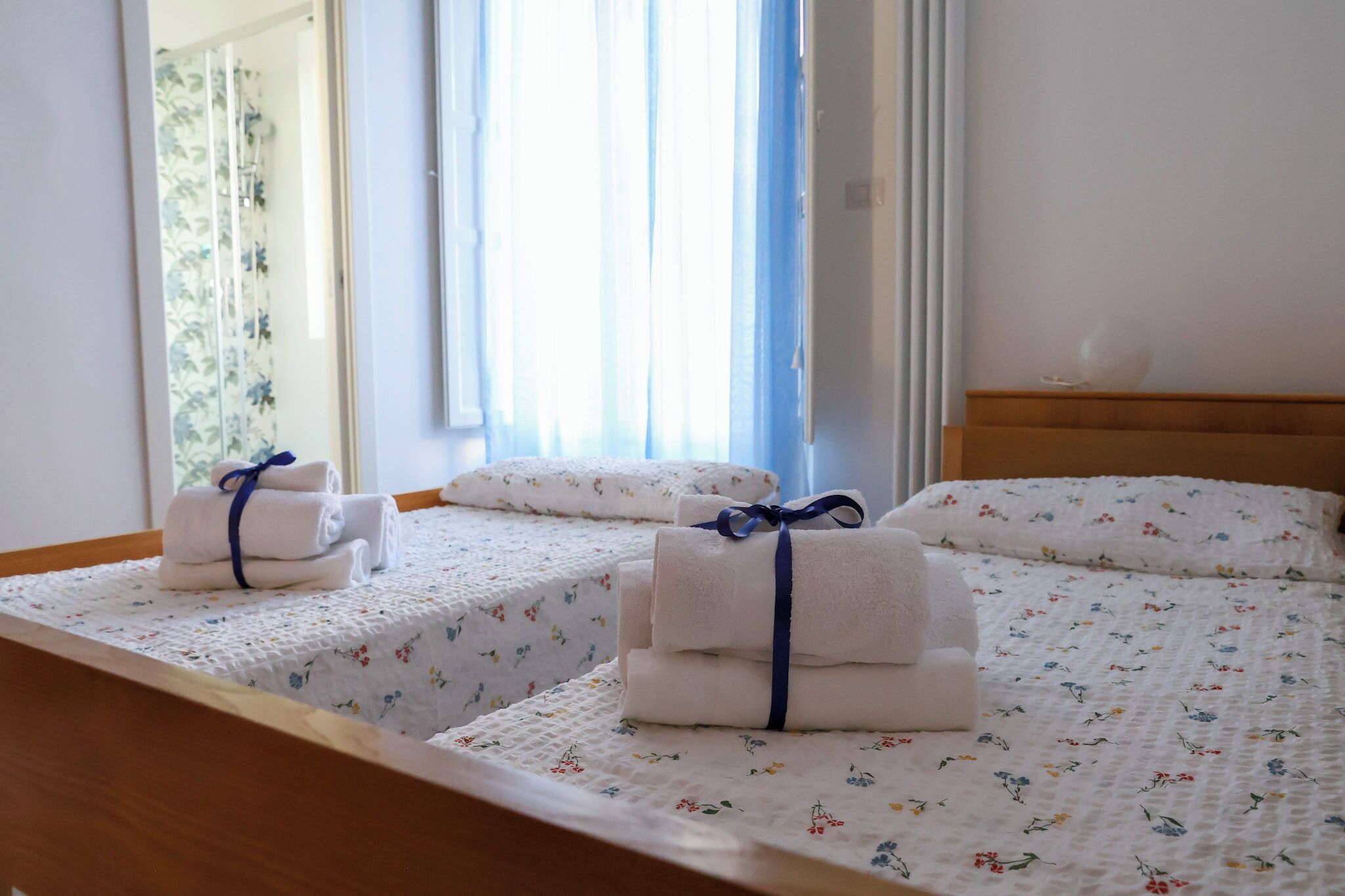 Une chambre d’hôtel avec deux lits, chacun recouvert d’une courtepointe à motifs floraux et agrémenté de serviettes soigneusement pliées, maintenues par un ruban bleu.