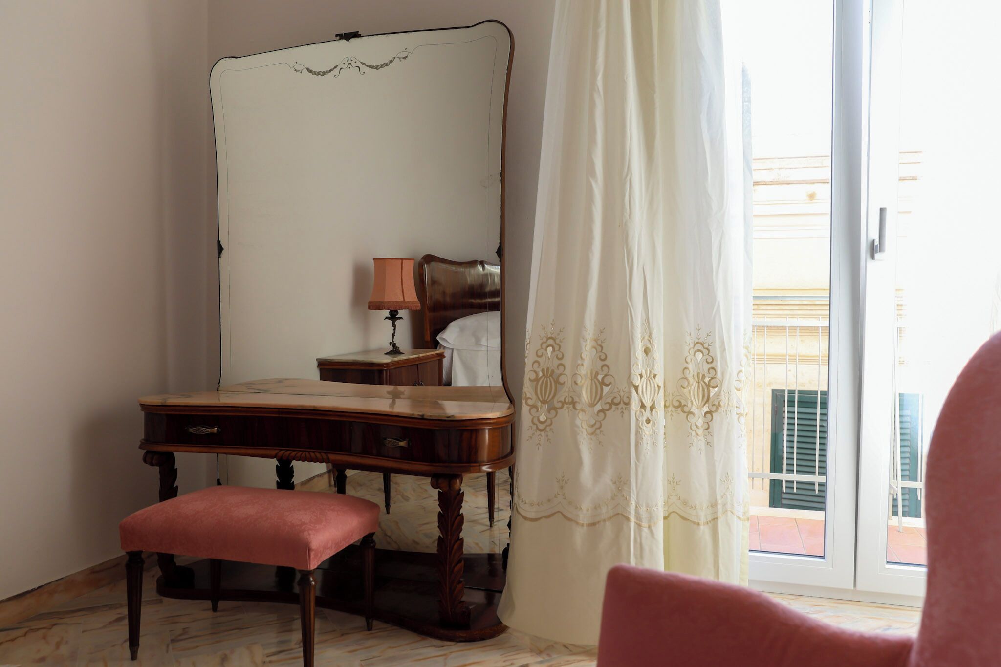 A room with a wooden desk, a chair, a mirror, a lamp, and a window with a curtain.