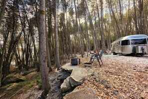 Un campeggio con una tenda, una sedia e un camper parcheggiati su terreno sterrato, circondato da alberi.