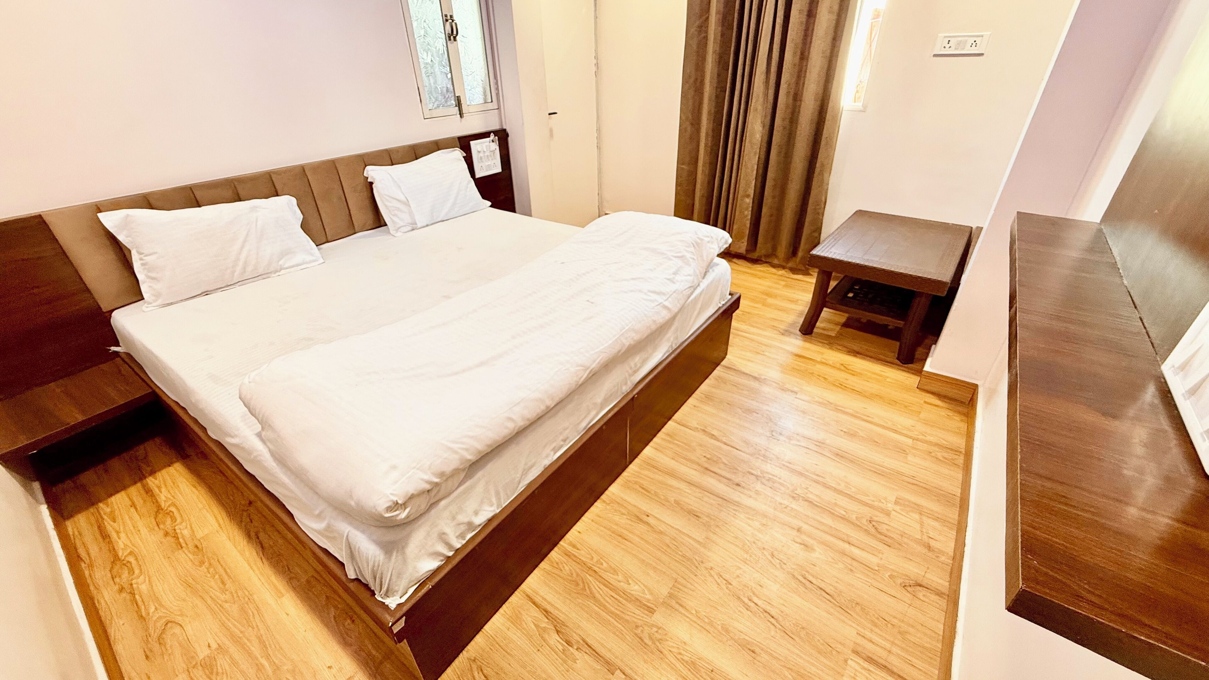 A neatly made bed with white linens in a hotel room with wooden flooring and a small bedside table.