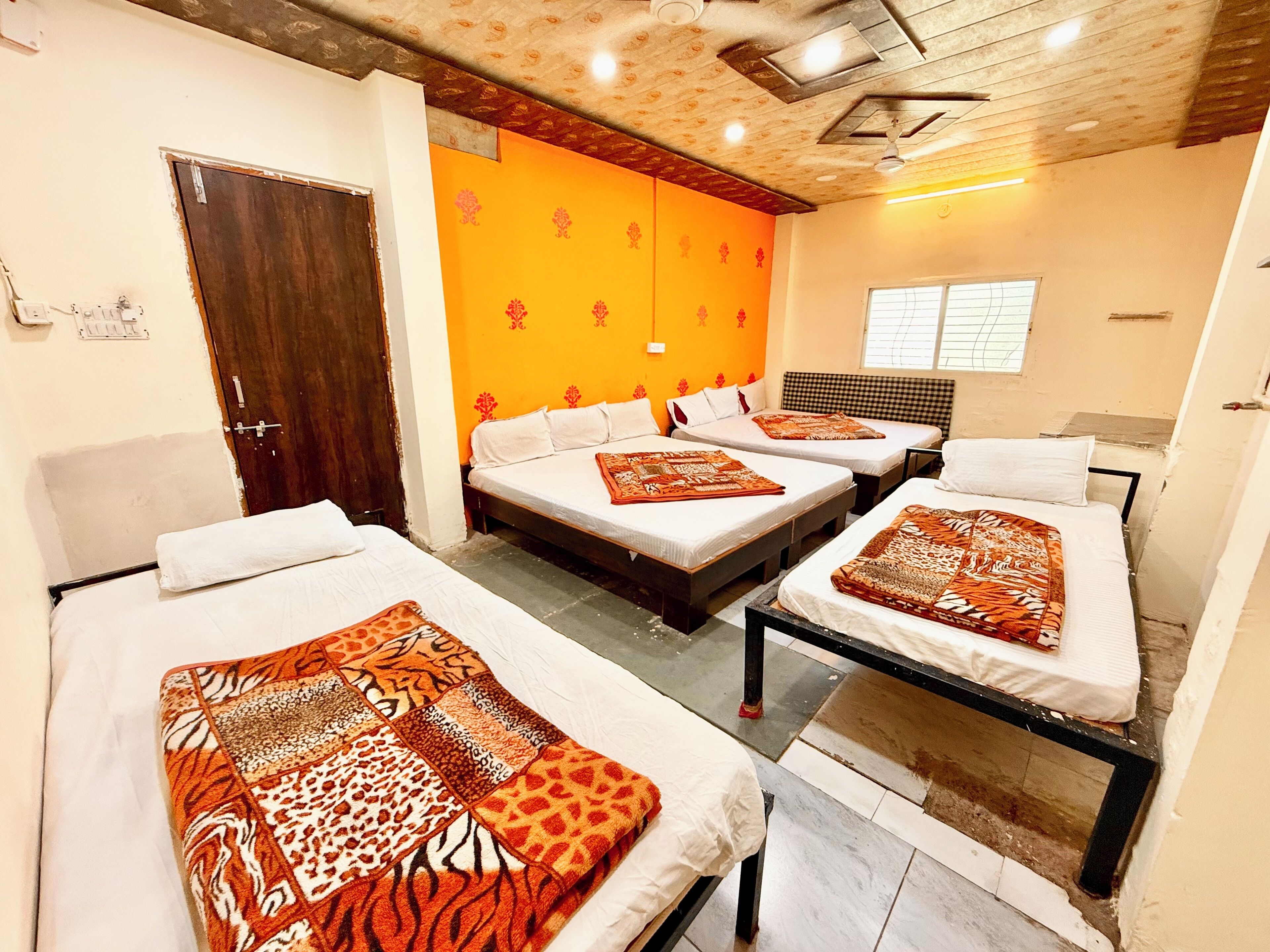 A hotel room with three beds, each with a patterned blanket. The room has an orange wall with floral patterns, a wooden ceiling, and a window with blinds.