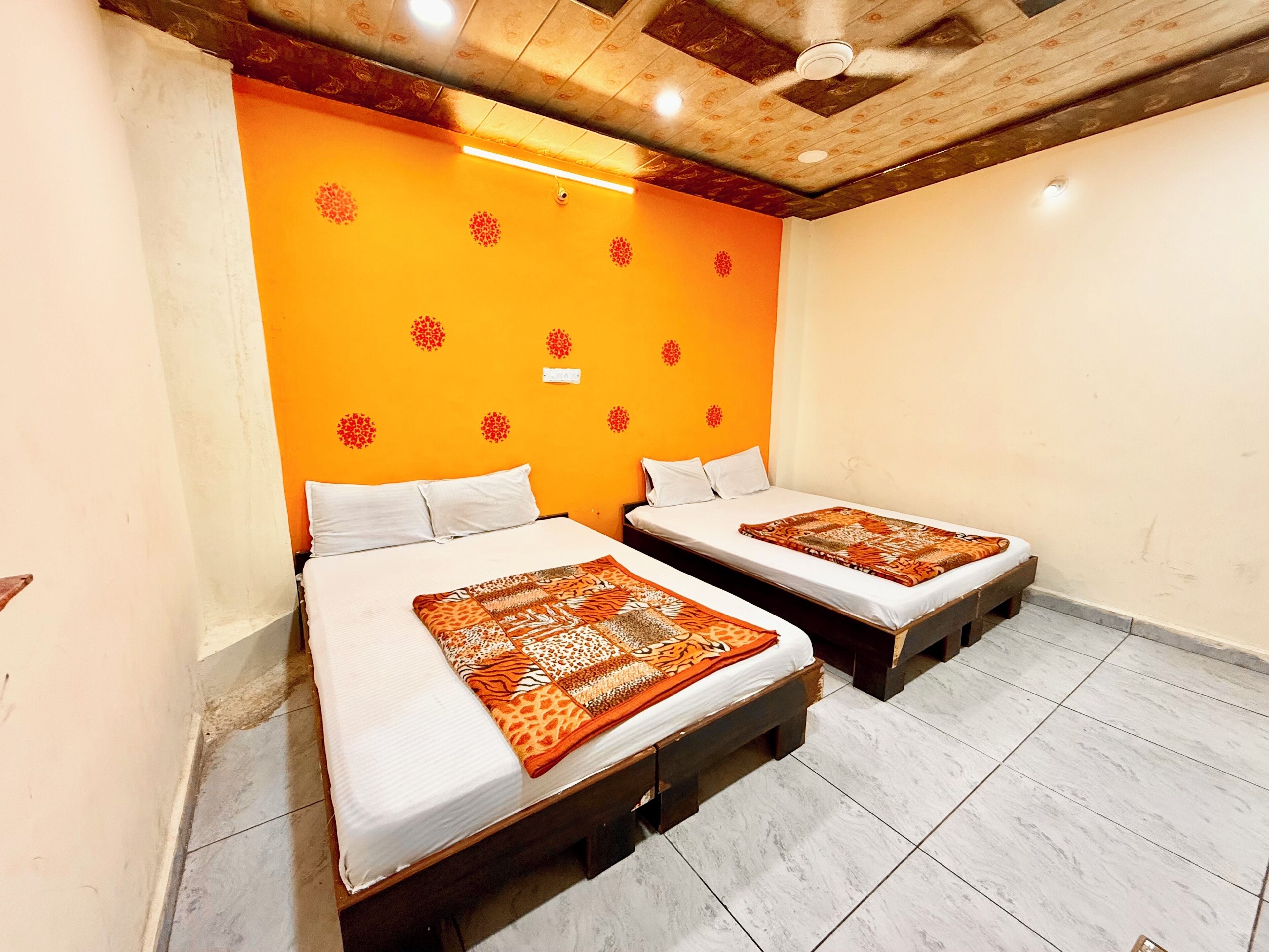 A room with two beds, each with a patterned bedspread, white pillows, and wooden headboards. The walls are orange with red floral decals, and the ceiling has exposed wooden beams.