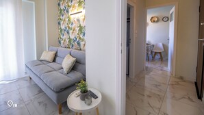 Una sala de estar moderna con un sofá gris, una mesita redonda con una planta y un control remoto, y una pared decorada con un estampado de hojas tropicales.