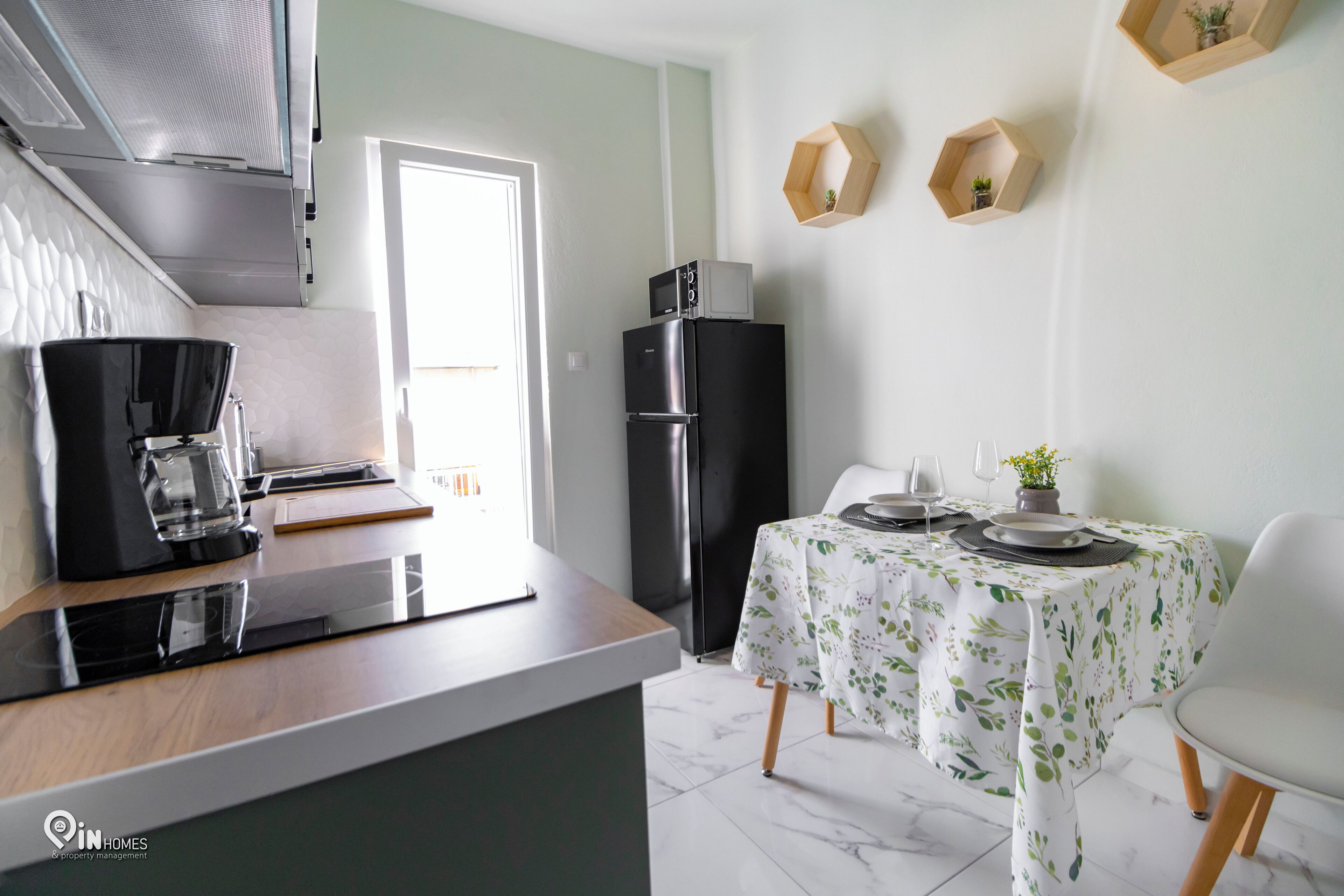 Eine moderne Küche mit Kaffeemaschine, Kühlschrank und Esstisch für zwei Personen, gedeckt mit einer blumigen Tischdecke.