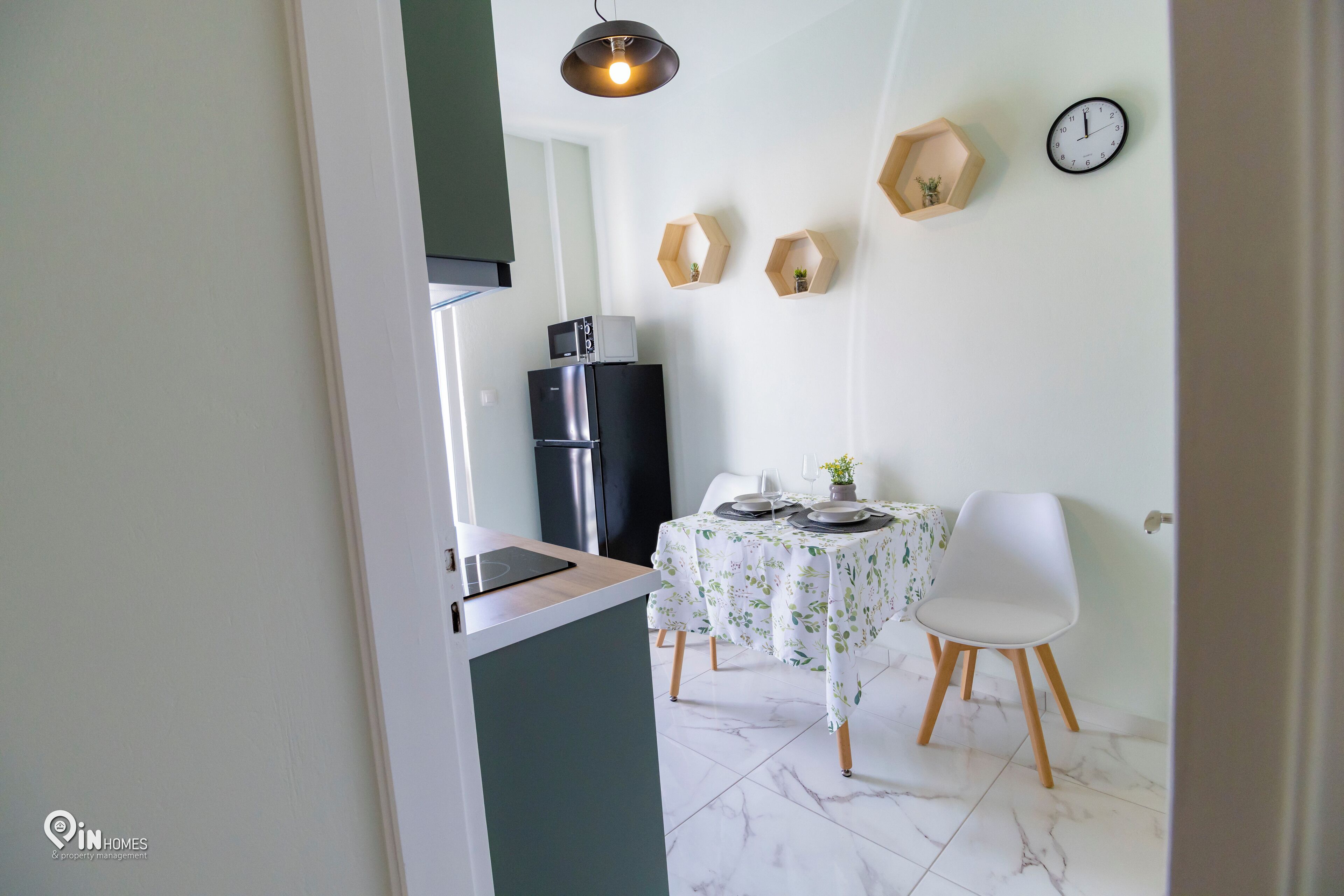 A modern kitchen with a built-in oven, a refrigerator, and a microwave. There is a dining table with a white tablecloth and two white chairs. A clock and hexagonal wall shelves are visible.