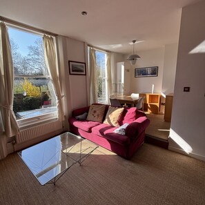 A living room with a red sofa, a glass coffee table, and a dining area in the background. - Brilliantly Located 1BDR Flat in West Kensington (Greater London)