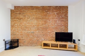 A room with a brick wall, a television on a wooden stand, and a black side table. - Brilliantly Located 1BDR Flat in West Kensington (Greater London)