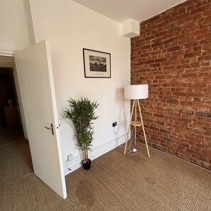 Une pièce avec une porte blanche, une plante en pot, un tableau encadré accroché au mur, un lampadaire blanc et un mur en briques.