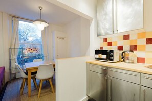 A compact kitchen with a microwave, toaster, and kettle on the counter, a dining table with chairs, and a view through a window. - Brilliantly Located 1BDR Flat in West Kensington (Greater London)