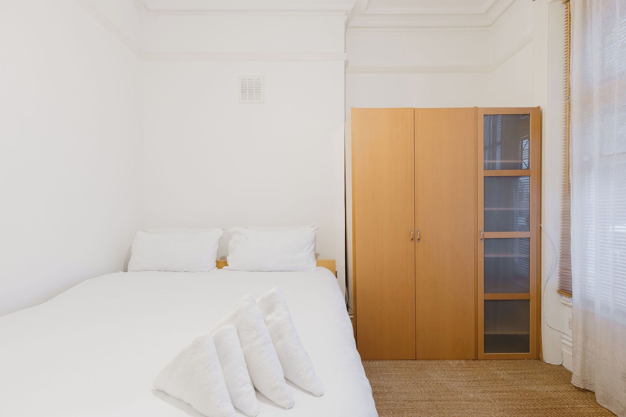 Un dormitorio con cama, ropa de cama blanca, un armario de madera y una ventana con persianas.