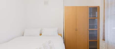 Une chambre à coucher avec un lit, du linge de lit blanc, une armoire en bois et une fenêtre avec des stores.