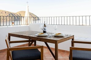 A wooden table with plates, wine glasses, and a bottle on a balcony with a view of hills and a lighthouse.