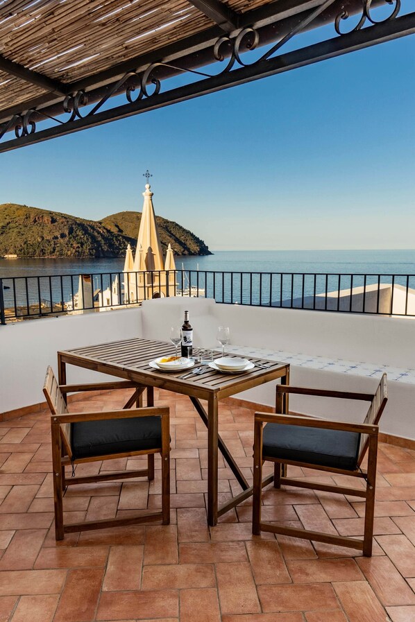 A terrace with a table and chairs, overlooking a coastal view with a church and hills.