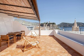 A terrace with a wooden table and chairs, a thatched roof, and a view of a coastal town.