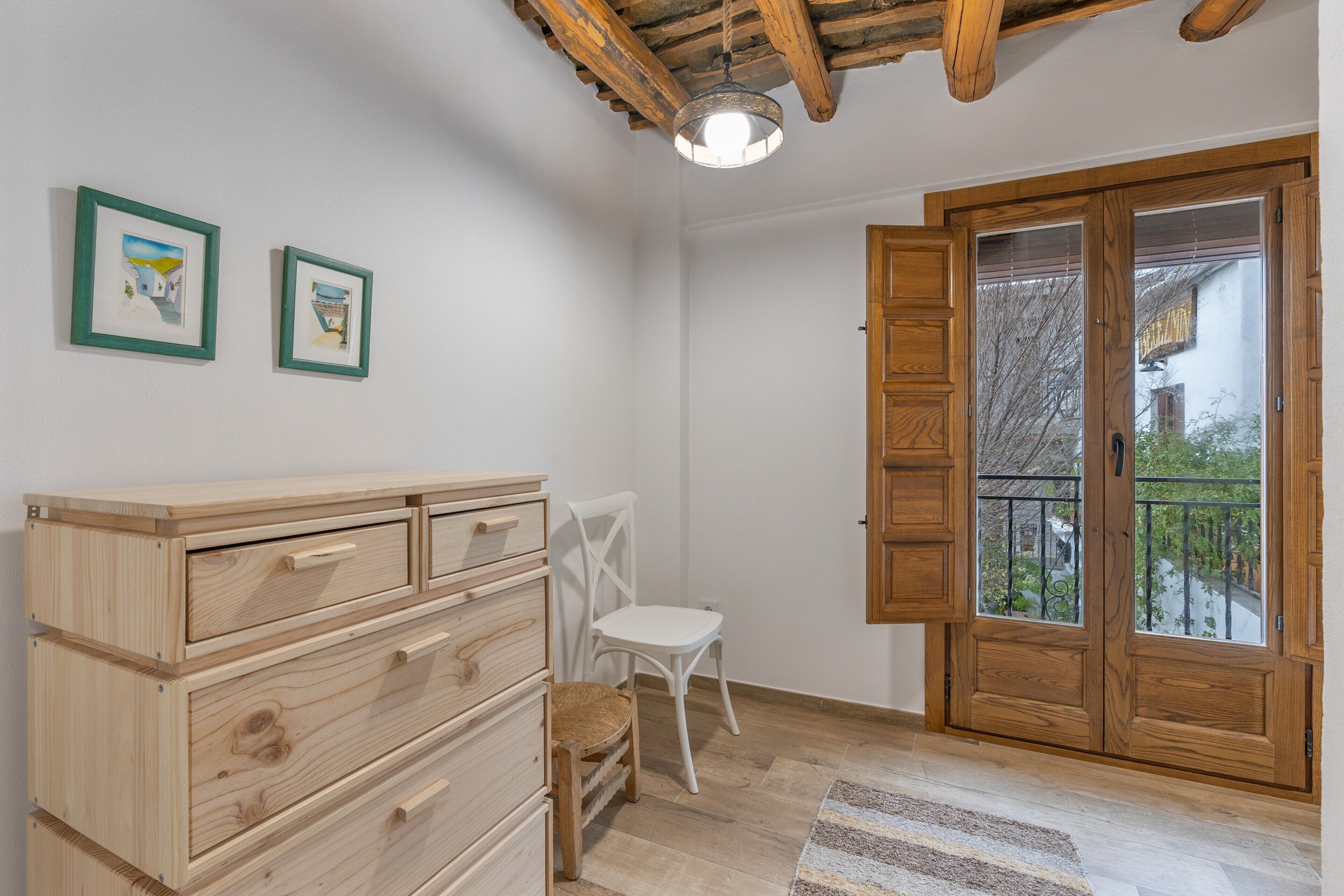 Una habitación con un aparador de madera, una silla blanca, una puerta de madera con balcón y dos cuadros en la pared.