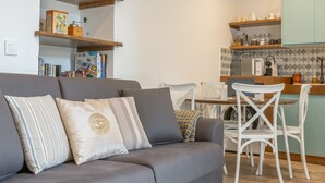 A modern living room with a grey sofa, white chairs, a kitchen area with wooden shelves, and a tiled backsplash. - Casa Pepa in Pampaneira – Charming Holiday Home in the Heart of La Alpujarra (Pampaneira)