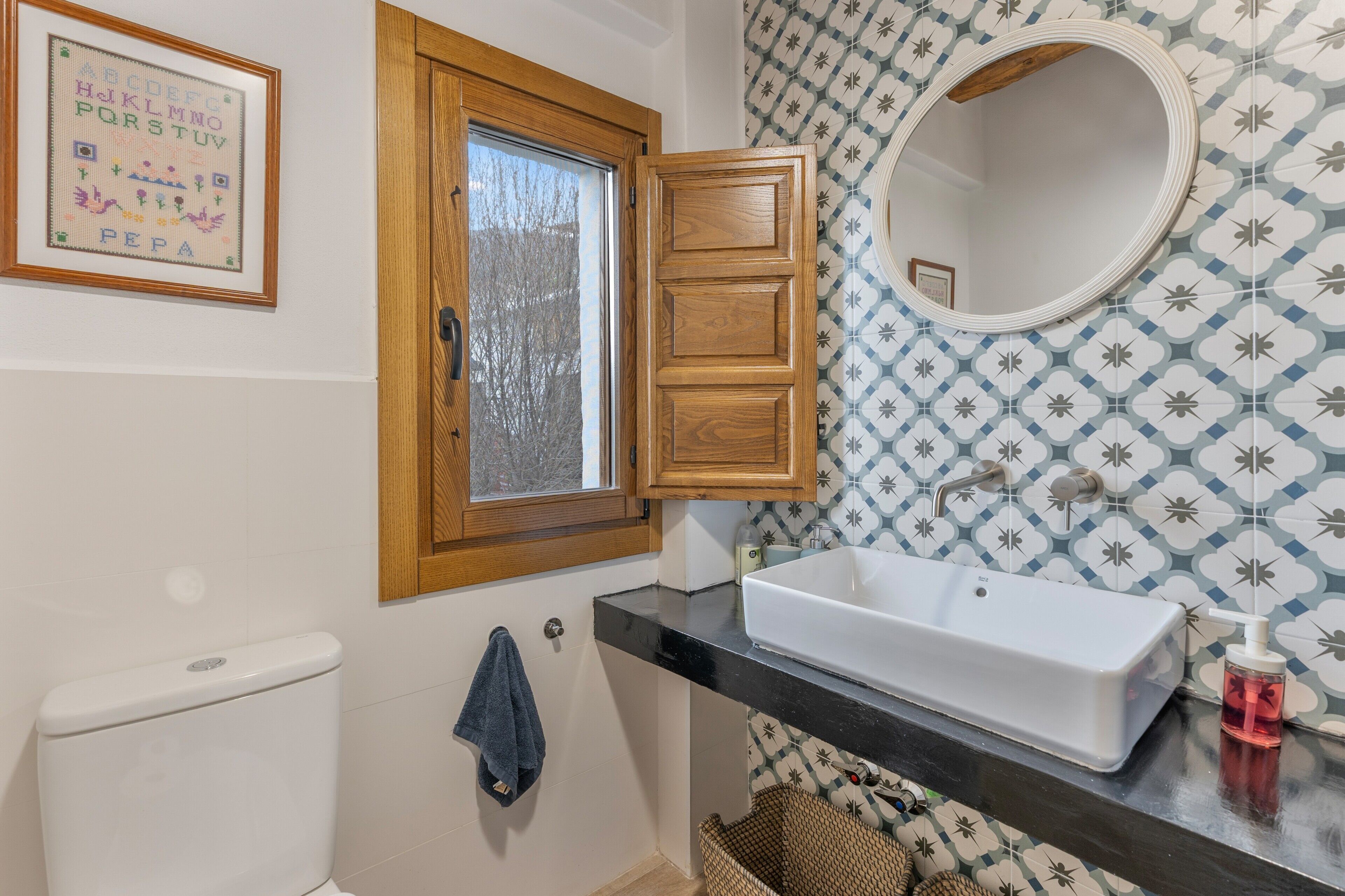 A bathroom with a white toilet, a sink with a mirror, a window, and patterned wallpaper.