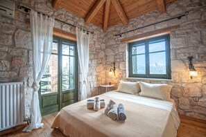 Une chambre aux murs de pierre et au plafond en bois, avec un lit recouvert de draps blancs, deux fenêtres ornées de rideaux et une table de chevet sur laquelle se trouve une lampe.