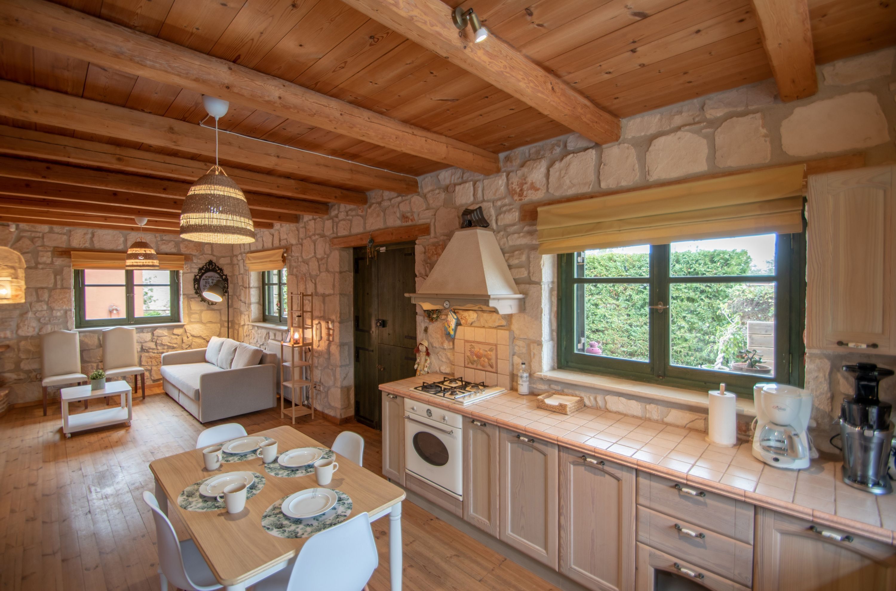 A rustic kitchen with stone walls, wooden beams, a dining area with a table set for four, and a window with a view of greenery.