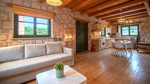 A cozy cabin interior with a stone wall, wooden ceiling, a sofa, a dining table, and a kitchen area. - Valory s Stone House (Zakynthos)