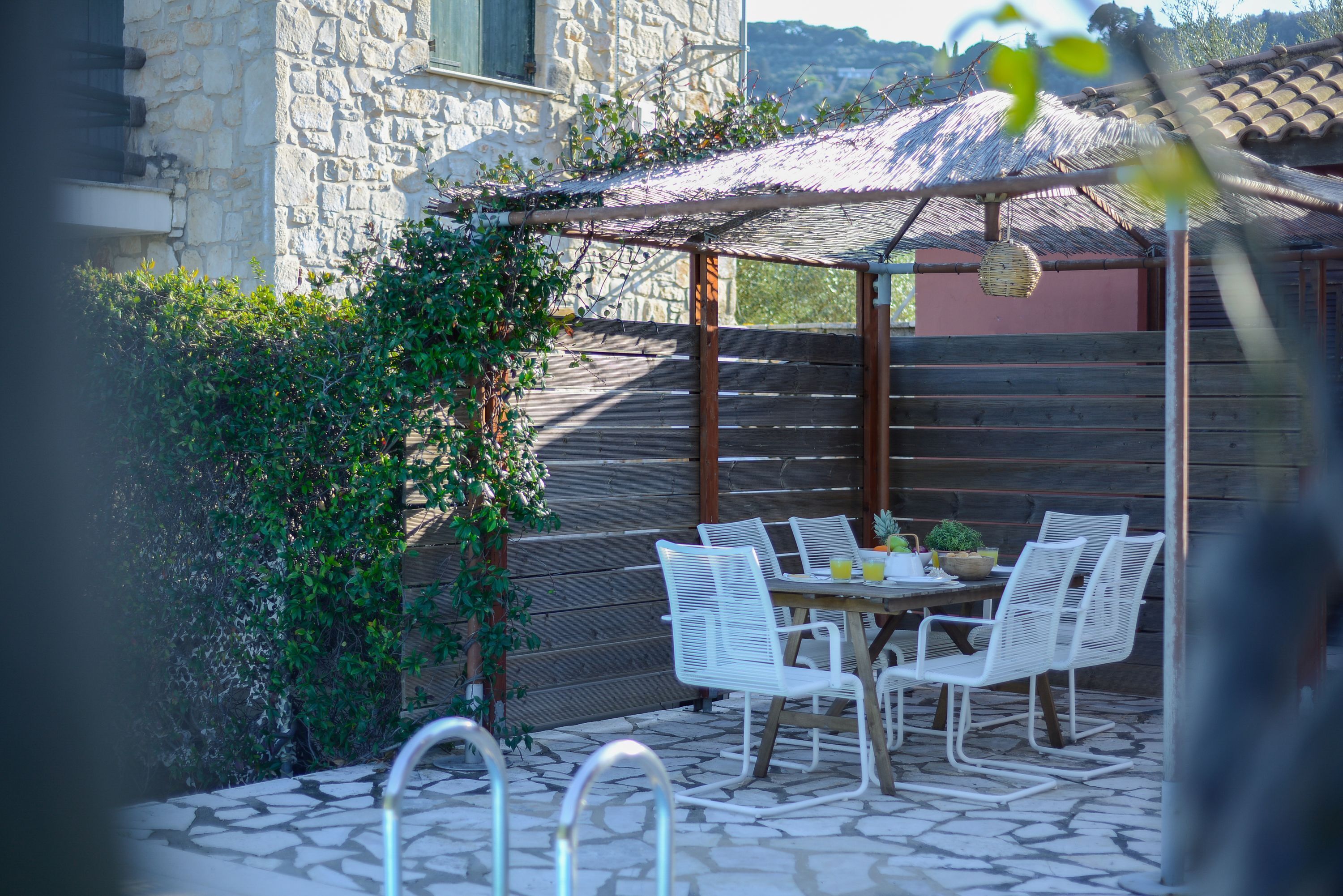 An outdoor patio with a stone floor, white metal chairs, a wooden table, and a pergola with a thatched roof.