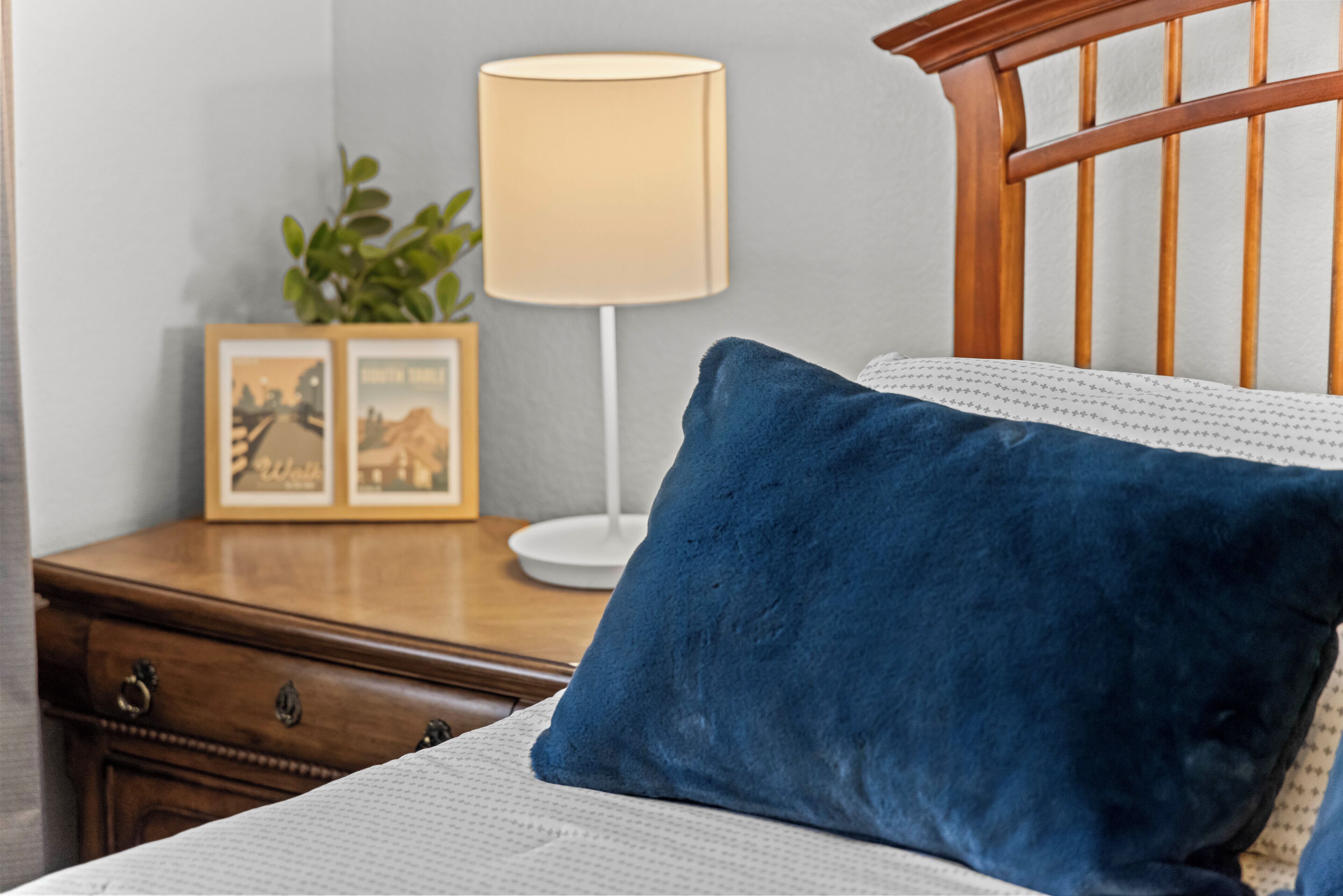 Ein ordentlich bezogenes Bett mit einem blauen Kopfkissen, ein Nachttisch aus Holz mit einer Lampe und gerahmte Bilder.