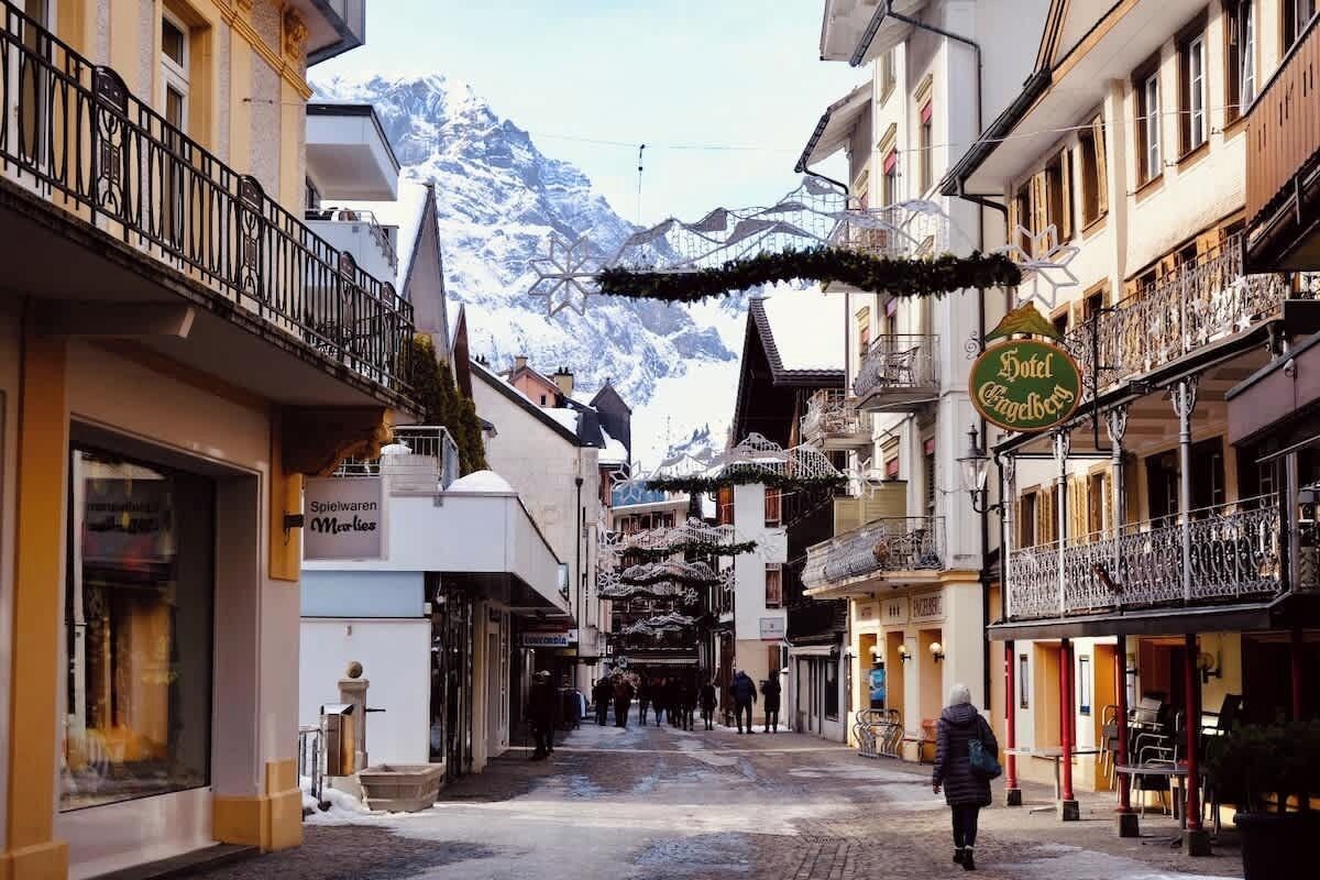 Une rue étroite bordée de bâtiments des deux côtés, une enseigne indiquant l’« hôtel Engelberg », et des montagnes enneigées en arrière-plan.