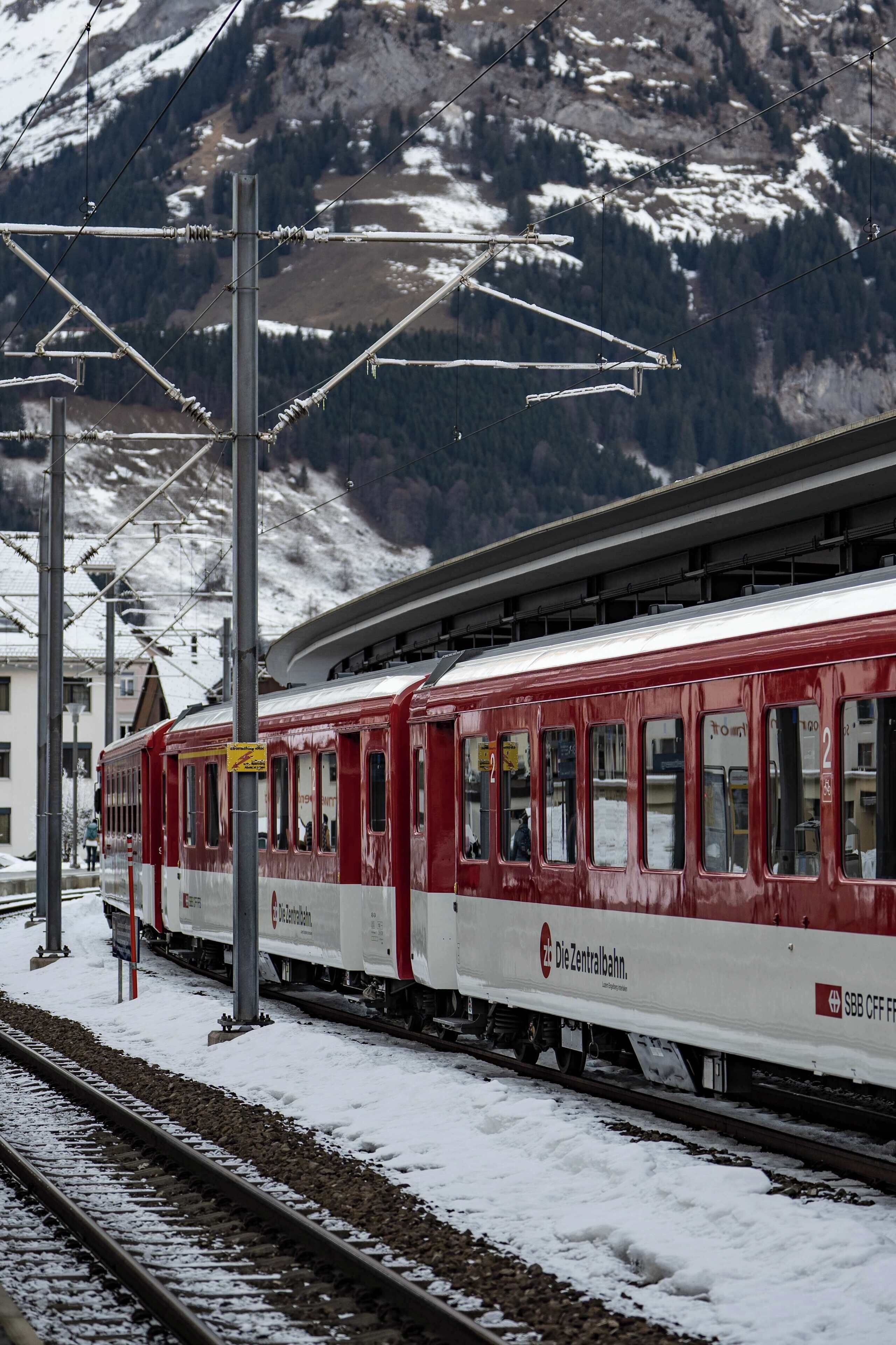 A red and white train at a station with snow on the ground and mountains in the background.
