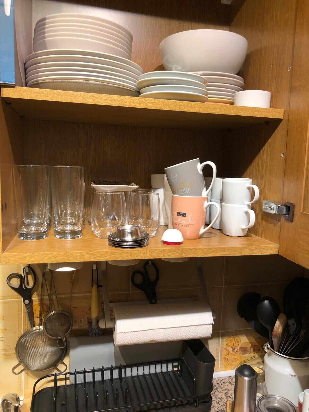 Ett kök med hyllor i trä där tallrikar, skålar och glas står. På köksbänken finns också köksredskap och en kaffebryggare.