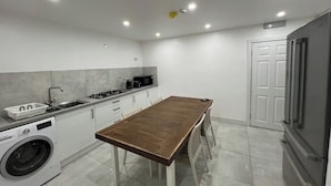 A compact kitchen with a wooden table, a washing machine, a microwave, and a refrigerator. - Luton City Retreat - Spacious 3 Bedroom Apartment (Luton)