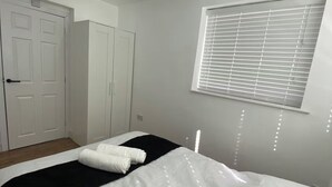 A bedroom with a bed, a window with blinds, a wardrobe, and a door. - Luton City Retreat - Spacious 3 Bedroom Apartment (Luton)