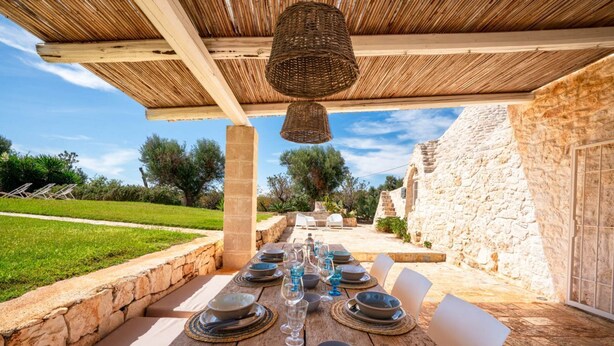 Un área de comedor al aire libre con una mesa de madera rústica dispuesta para cuatro personas, rodeada de muros de piedra y un cielo despejado.