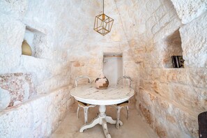 Ein Zimmer mit Steinwänden, einem runden Tisch, zwei Stühlen, einer Pendelleuchte und einem kleinen Bücherregal.
