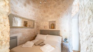 Un dormitorio con paredes de piedra, una cama, almohadas, una mesita de noche con una lámpara y un espejo.