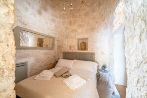 Ein Schlafzimmer mit Steinwänden, einem Bett, Kissen, einem Nachttisch mit Lampe und einem Spiegel.
