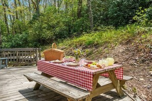 Ein Picknick mit karierter Tischdecke, einem Korb und Lebensmitteln auf einer Holzterrasse, umgeben von Bäumen.