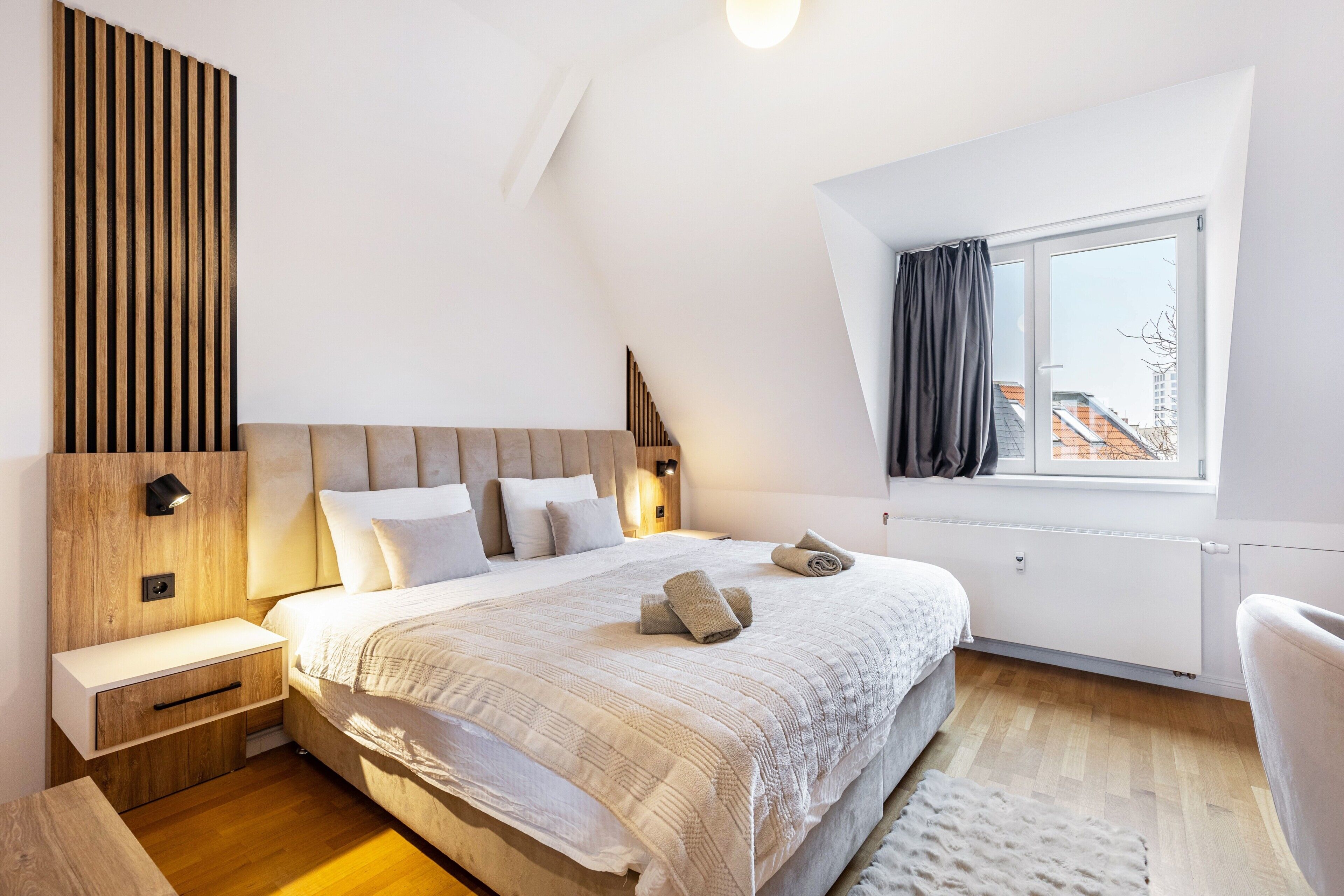 A modern bedroom with a sloped ceiling, a large bed with a headboard, bedside tables, a nightstand, a window with curtains, and a view of buildings outside.