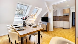 Un coin repas avec une table en bois, des chaises blanches et un vase rempli de fleurs. On aperçoit une télévision et une cuisine au fond.