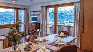 Ein Essbereich mit einem Holztisch, gedeckt für zwei Personen, großen Fenstern mit Bergblick, einem Fernseher auf einer Holzkommode und einer Tannenzweiganordnung.