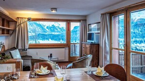 Ein Essbereich mit einem Tisch für vier Personen, große Fenster mit Blick auf die Berge, ein Fernseher auf einem Holzschrank und eine gemütliche Sitzecke mit einer Couch und Sesseln.