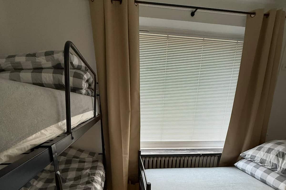 A dormitory room with bunk beds, a window with blinds, and beige curtains.