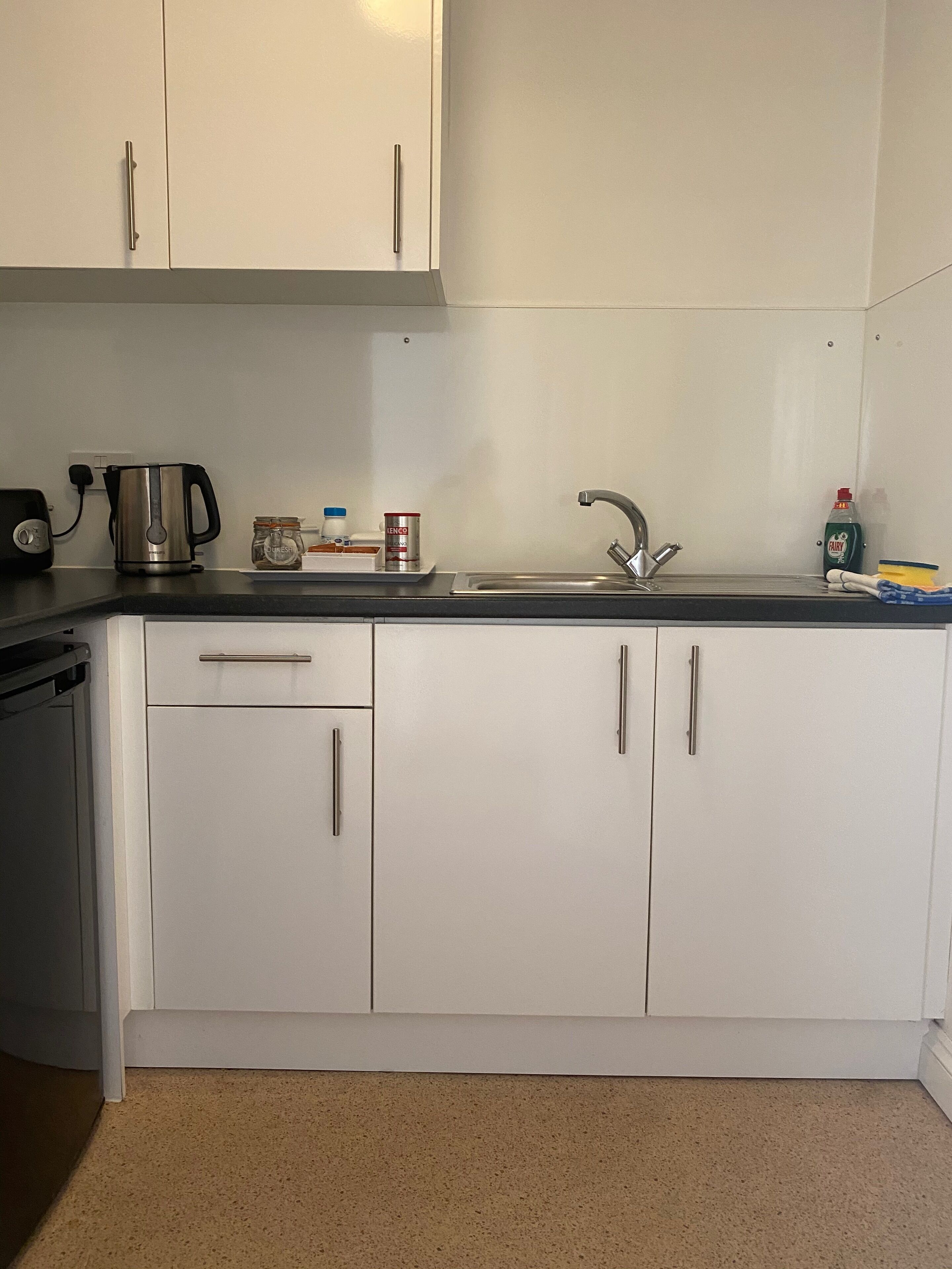 A kitchen with white cabinets, a black countertop, a kettle, a jar, a can, a bottle of dish soap, and a toaster.