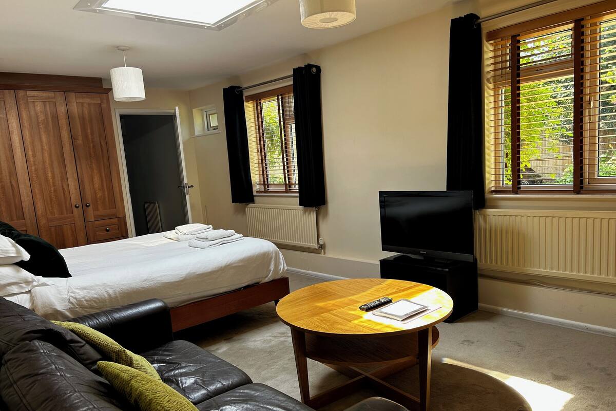 A hotel room with a bed, a wooden table, a leather sofa, a TV, and windows with curtains.
