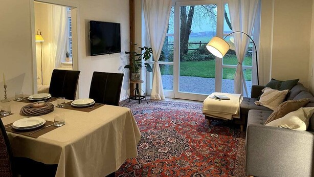A dining area with a table set for four, a sofa, and a TV mounted on the wall. - Landhaus Baumberge - Cozy vacation home & sauna (Billerbeck)