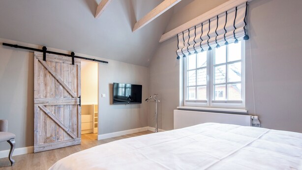 A bedroom with a large bed, a sliding barn door, a television, and a window with curtains. - House SyltZauber - Sylt (Sylt)
