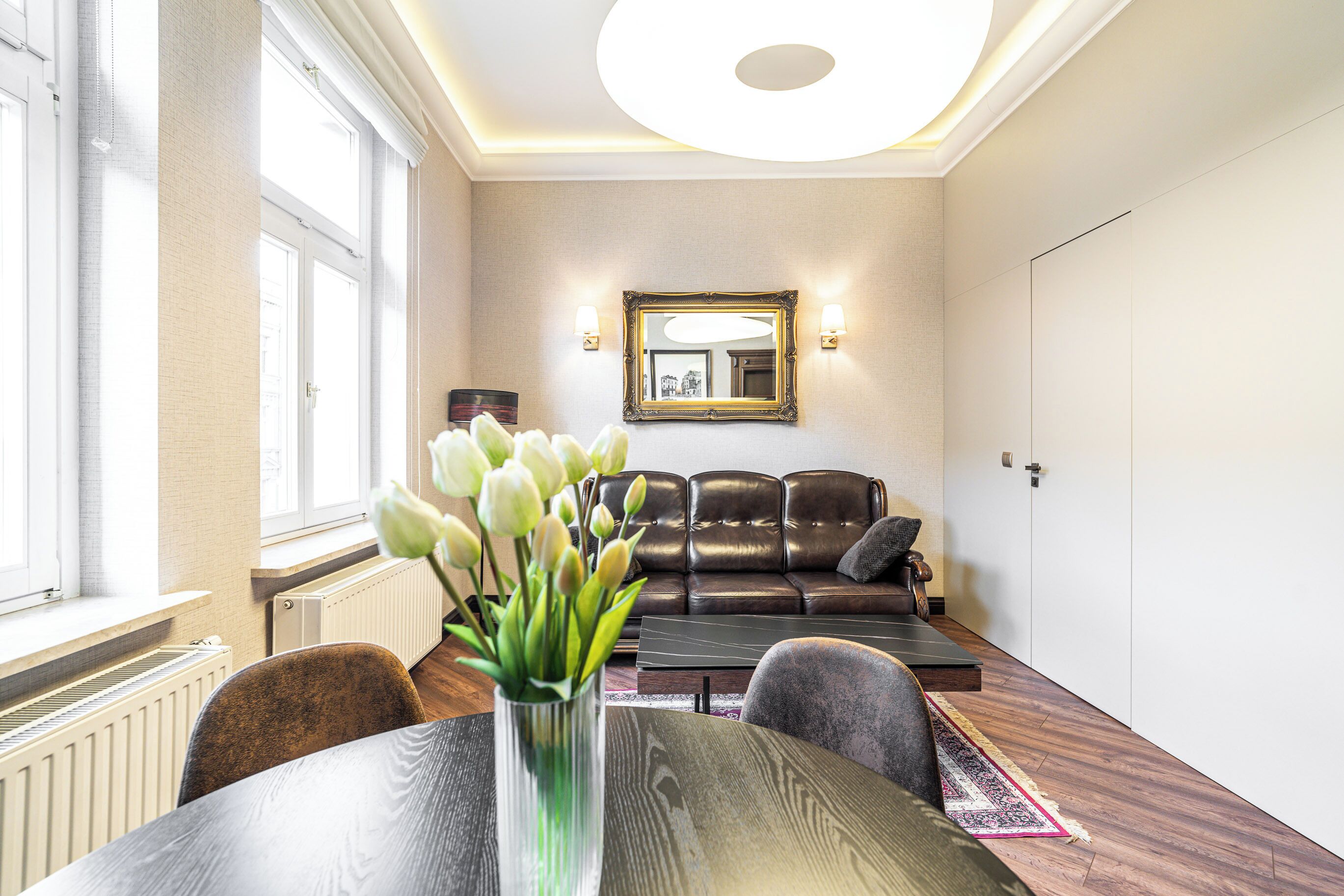 Ein modernes Wohnzimmer mit einem Ledersofa, einem Esstisch mit Blumenvase und einem großen Spiegel an der Wand.