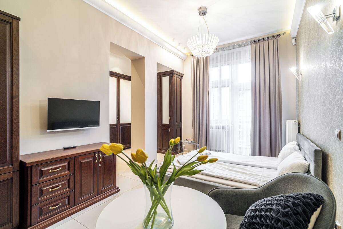 A hotel room with a bed, a TV, a desk with a vase of flowers, and a view of the city through large windows.
