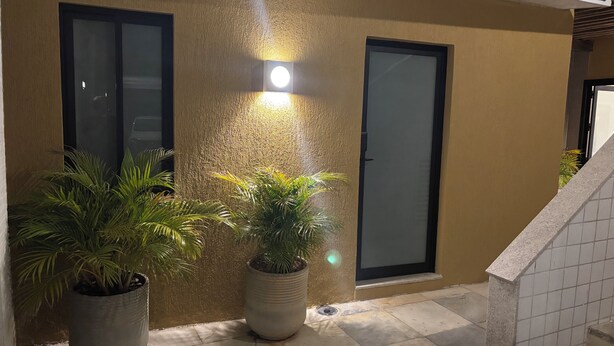 Fachada moderna de edificio con paredes texturizadas, dos puertas de vidrio y una lámpara empotrada iluminada. En primer plano, hay dos plantas grandes en macetas.