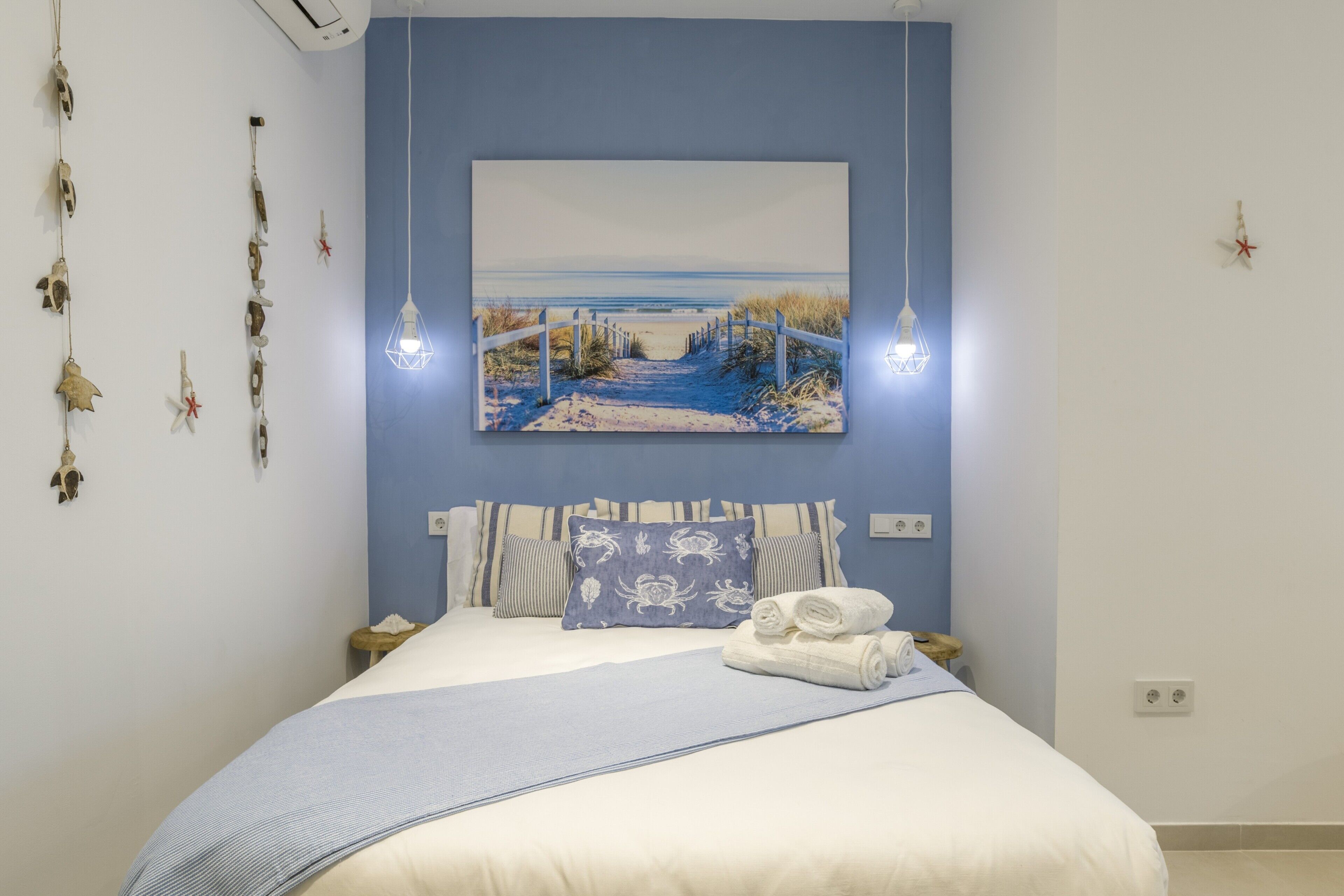 Ein ordentlich bezogenes Bett mit blauer Decke, ein Strandbild darüber und eine Hängeleuchte.