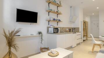 Una cocina moderna con armarios blancos, un televisor de pantalla plana, repisas de madera con utensilios de cocina, una zona de comedor con una mesa y sillas blancas, y una planta pequeña.